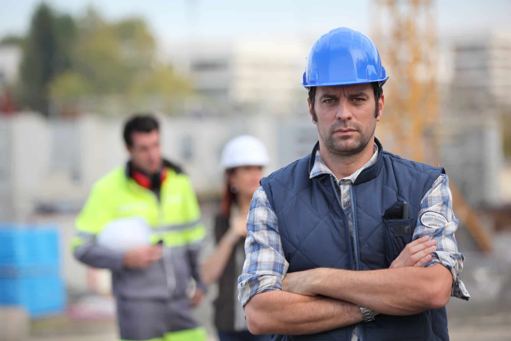 Electrician with folded arms and blue hard hat. Two electricians in background.